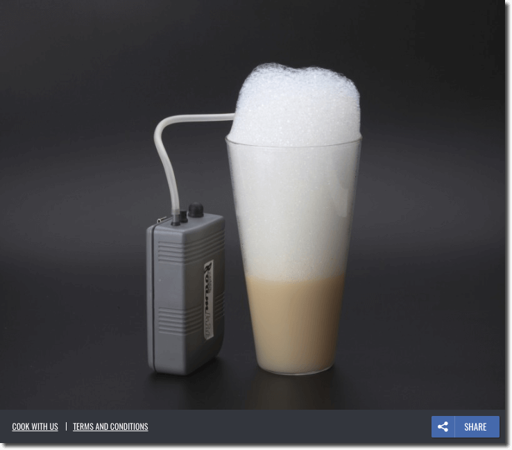 The image shows a foaming tool: a grey box with a rubber pipe leading into a glass of coffee, which is filled to overflowing with foam. The background is a deep, minimalist grey. The only text is 3 small buttons at the bottom of the image: "cook with us", "terms and conditions", and "share".